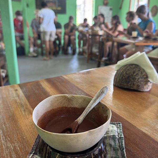 A handmade chocolate drink served in traditional Maya bowls from Ajaw Chocolate, a family-run business preserving the art of traditional Maya chocolate-making. Background: Students learning to add local honey and cayenne to their chocolate drink as is customary in ancient Maya chocolate-making. [photo courtesy of Erin Wahler]
