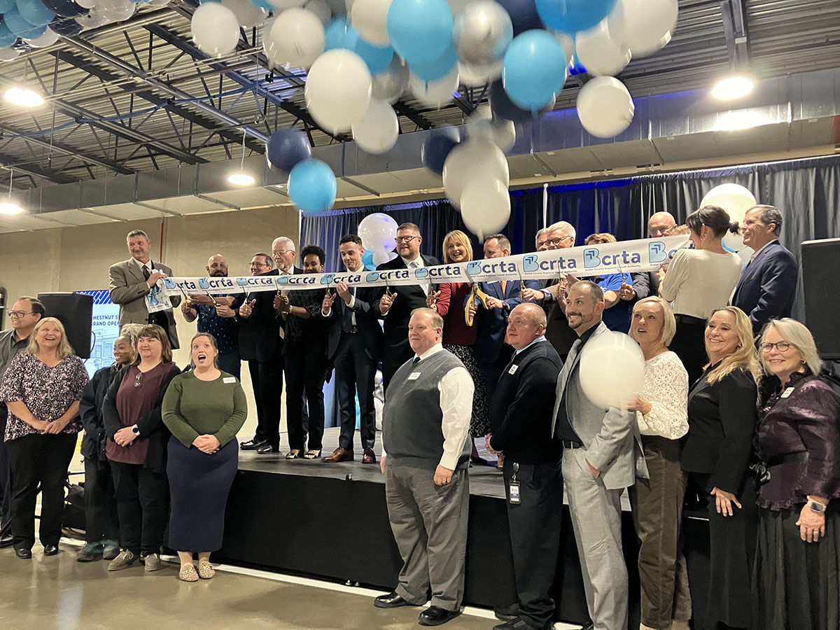 a group of dozens of people involved with the Chestnut Street Station stand on the podium and cut a large ribbon, with blue and white balloons overhead 