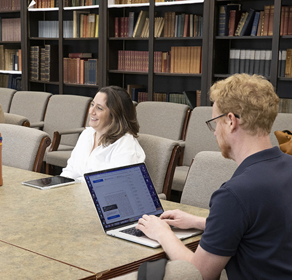 Two people sit a large table , one has a laptop open; a wall-lined bookcase filled with books is behind them